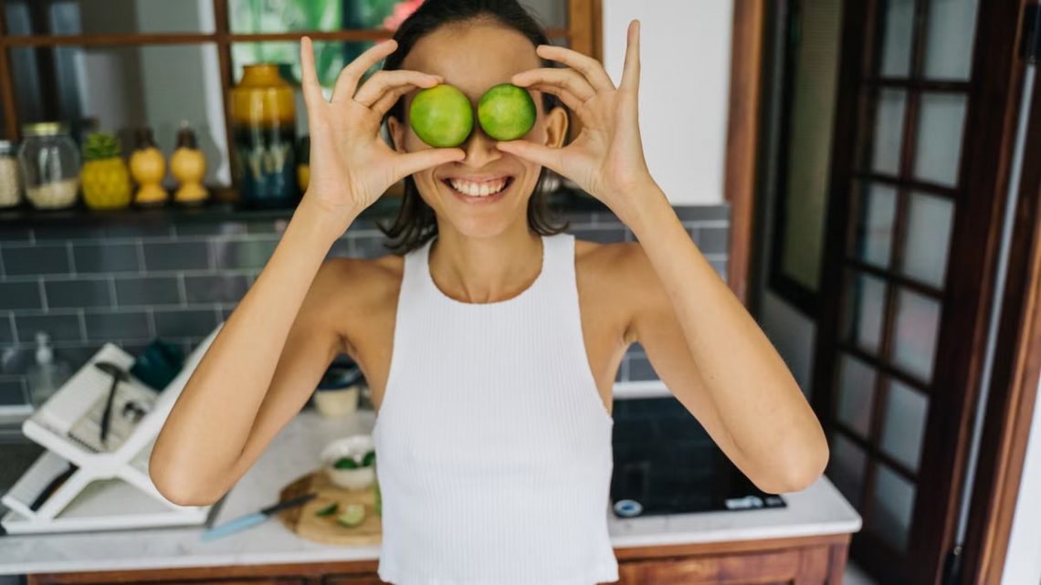 Smiling young woman playfully holding limes over her eyes in a bright kitchen, capturing a fun and positive moment.
