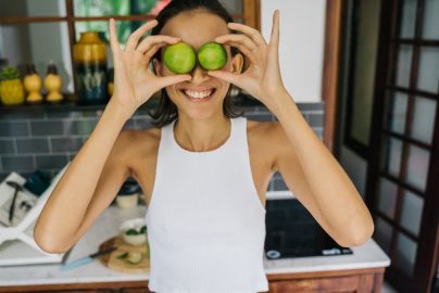 Smiling young woman playfully holding limes over her eyes in a bright kitchen, capturing a fun and positive moment.